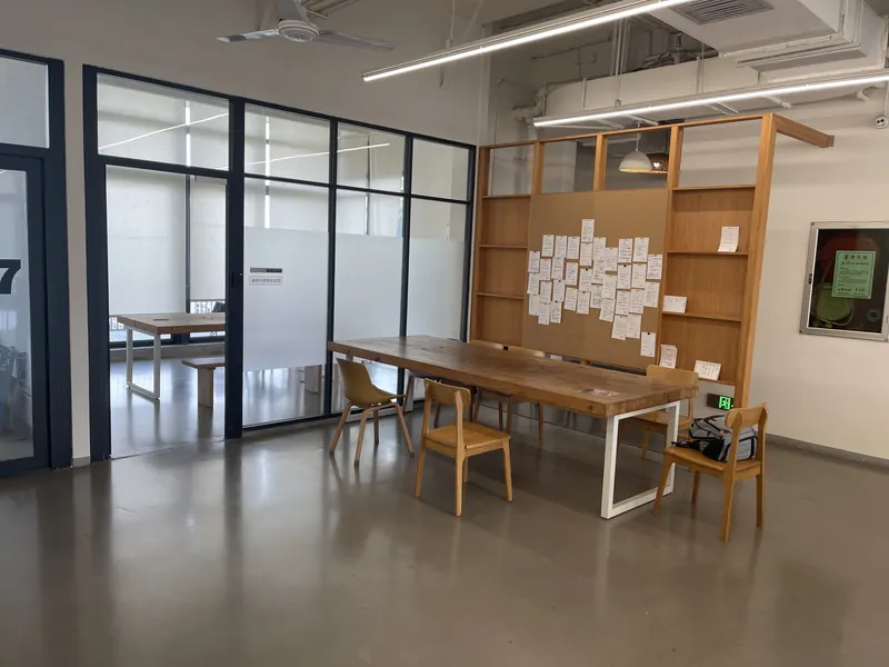 Meeting table with wooden chairs beside a cork notice board and a glass-walled meeting room