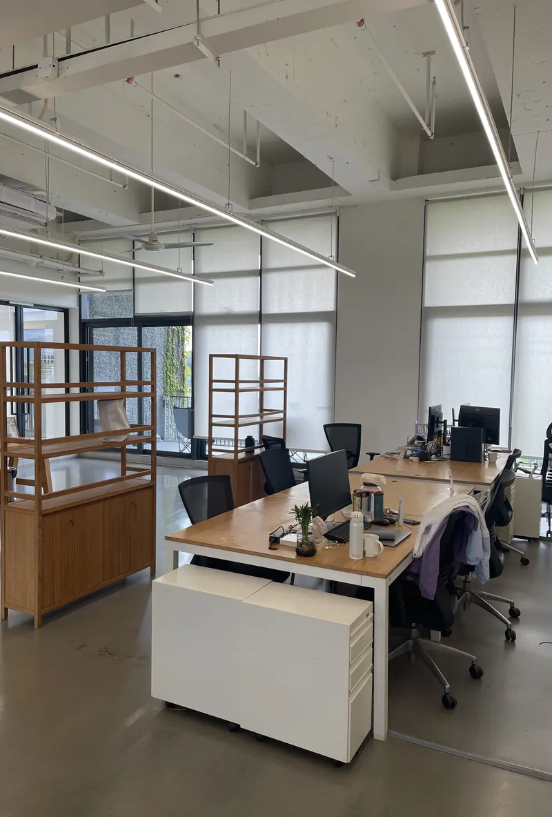 Open-plan office with desks, monitors and wooden shelving under tall windows